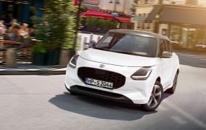 Weißer Suzuki Swift 1.2 DUALJET HYBRID Comfort+ vor City-Setting und Restaurant-Außenbereich in Front-Seitenansicht