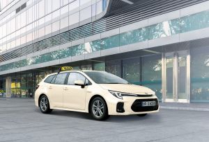 Beiger Suzuki Swace 1.8 HYBRID CVT Comfort+ mit Taxischild vor Glasfront eines Gebäudes in Front-Seitenansicht